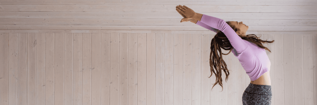 A woman with dark long hair wearing a pink top and grey tights is stretched up and back with her arms in the air